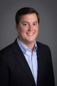 A man in a dark suit and blue shirt smiling warmly at the camera.