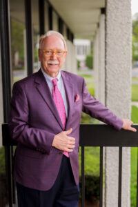 A man with white hair and glasses wearing a vibrant purple suit jacket, smiling confidently while standing against a neutral background. Ed Smith